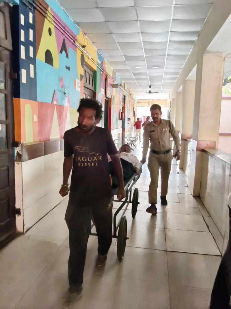 रंगीन दीवार वाले अस्पताल गलियारे में स्ट्रेचर खींचता आदमी, पीछे खाकी वर्दी में पुलिसकर्मी